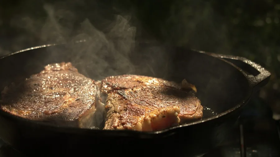 Close-up of sizzling steak being seared in a cast iron skillet on a stovetop with steam rising
