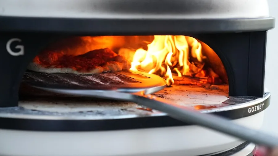 Family gathered around glowing outdoor pizza oven at sunset, flames visible through opening